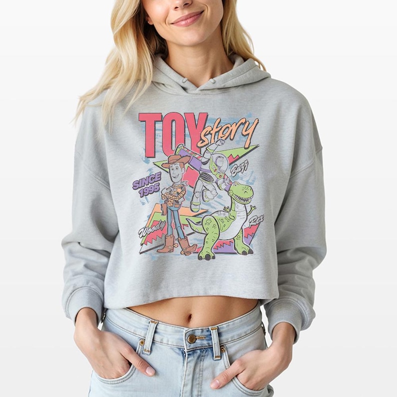 May include: A light grey cropped hoodie featuring a colourful graphic print of Toy Story characters Woody, Buzz Lightyear, and Rex. The design includes the text Toy Story, Since 1995, Woody, Buzz, and Rex, set against a background of geometric shapes.