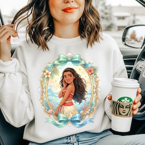 May include: White sweatshirt featuring a colorful illustration of a woman in a tropical setting, framed by a gold border, bows, and floral accents. The woman is depicted with long, dark hair and a red and white outfit. A Starbucks coffee cup is held in the hand.