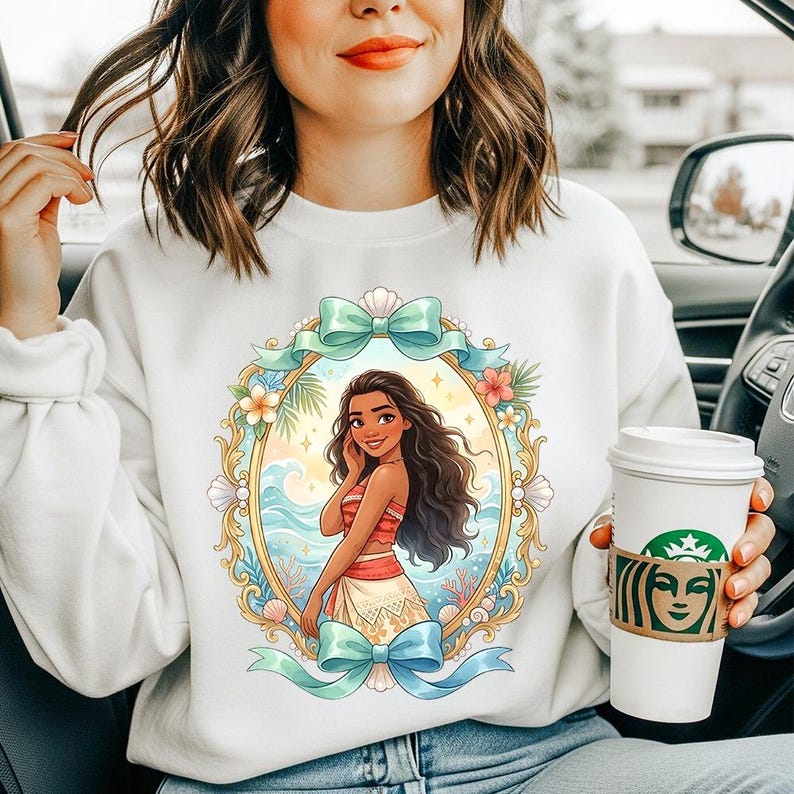 May include: White sweatshirt featuring a colorful illustration of a woman in a tropical setting, framed by a gold border, bows, and floral accents. The woman is depicted with long, dark hair and a red and white outfit. A Starbucks coffee cup is held in the hand.