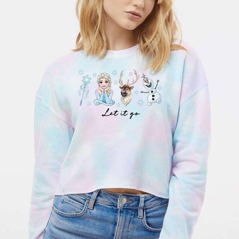 May include: A tie-dye cropped sweatshirt with a light blue, pink, and white color scheme. The front features a graphic of Elsa, Olaf, Sven, and a snowflake wand, with the text "Let it go" in black script.