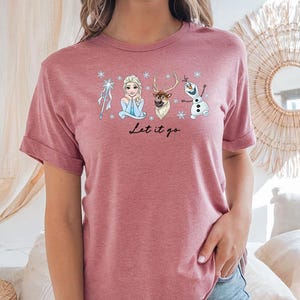 May include: A dusty rose t-shirt featuring a graphic of characters from the movie Frozen, including Elsa, Olaf, and Sven, with the text "Let it go" in a script font. The shirt has rolled-up sleeves and is made of a soft material.