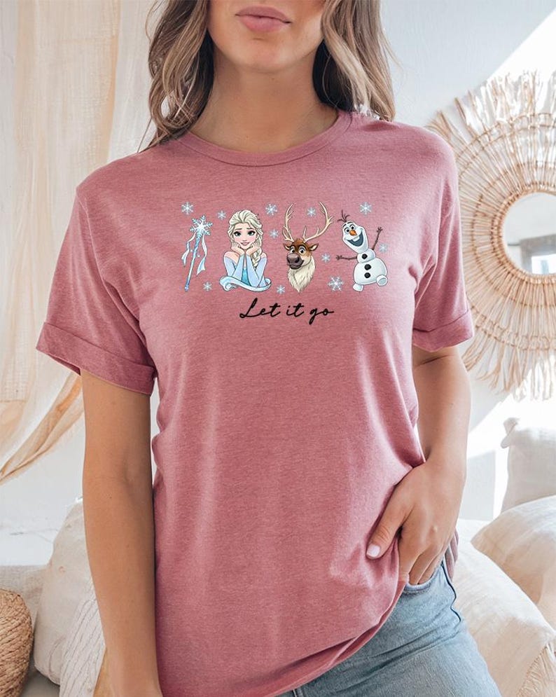 May include: A dusty rose t-shirt featuring a graphic of characters from the movie Frozen, including Elsa, Olaf, and Sven, with the text "Let it go" in a script font. The shirt has rolled-up sleeves and is made of a soft material.