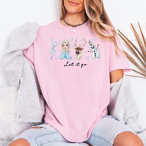 May include: Pale pink t-shirt with a graphic of Elsa, Olaf, and Sven from Frozen, with snowflakes and a wand. The words "Let it go" are written in black script. The shirt is made of a soft material.