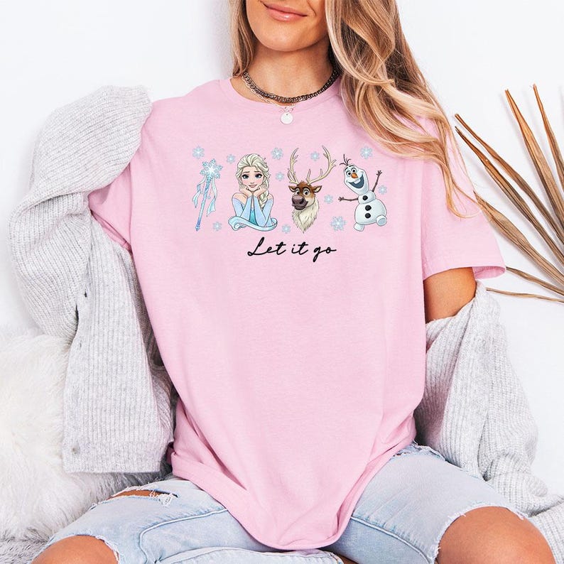 May include: Pale pink t-shirt with a graphic of Elsa, Olaf, and Sven from Frozen, with snowflakes and a wand. The words "Let it go" are written in black script. The shirt is made of a soft material.