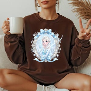 K&ouml;nnte beinhalten: Ein braunes Sweatshirt mit einer gerahmten Illustration einer Figur mit blonden Haaren und einem blauen Kleid. Der Rahmen ist mit Schneeflocken und blauen Schleifen verziert. Das Sweatshirt hat eine einfarbige braune Farbe.