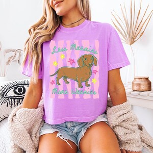 May include: A lavender short-sleeve t-shirt featuring a graphic of a brown dachshund dog centred between pink wavy vertical stripes. The text "Less Meanies More Weenies" is printed in green, surrounded by small yellow and pink star illustrations.