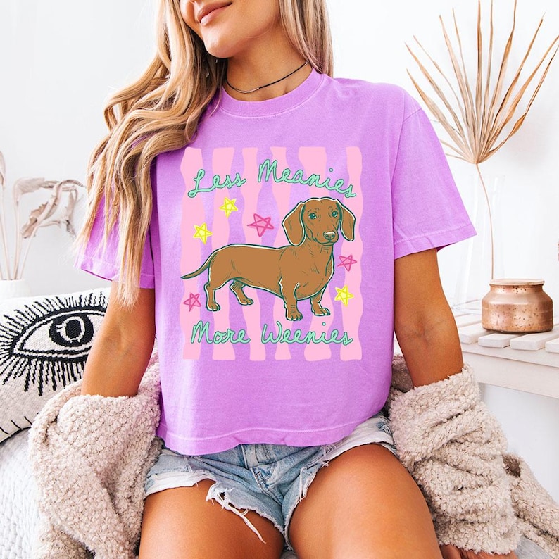May include: A lavender short-sleeve t-shirt featuring a graphic of a brown dachshund dog centred between pink wavy vertical stripes. The text "Less Meanies More Weenies" is printed in green, surrounded by small yellow and pink star illustrations.