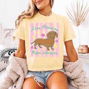 May include: A pale yellow short-sleeve t-shirt featuring a brown dachshund illustration centred on a pink wavy background. The text reads Less Meanies More Weenies in teal script, surrounded by small neon yellow and pink star outlines.