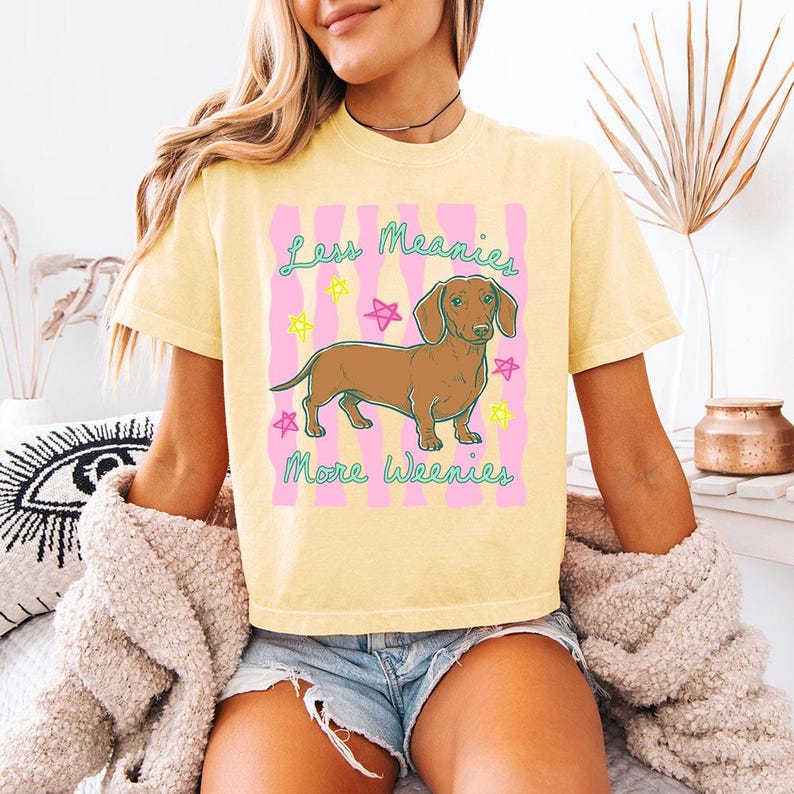 May include: A pale yellow short-sleeve t-shirt featuring a brown dachshund illustration centred on a pink wavy background. The text reads Less Meanies More Weenies in teal script, surrounded by small neon yellow and pink star outlines.