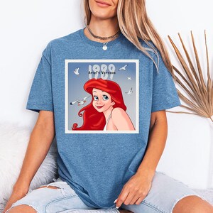 May include: Blue t-shirt featuring a square graphic of Ariel from The Little Mermaid, with the text "1989 Ariel's Version". The graphic includes Ariel's portrait, seagulls, and a blue background. The shirt is a solid blue color.