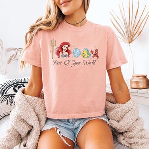 May include: A light pink t-shirt featuring a graphic of Ariel, Flounder, Sebastian, and a seashell. The phrase "Part Of Your World" is written below the characters. The shirt has short sleeves.