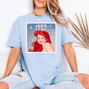 May include: Light blue t-shirt featuring a square graphic of Ariel from The Little Mermaid, with the text "1989 Ariel's Version". The graphic shows Ariel with red hair and seagulls in the background. The shirt is a casual style.