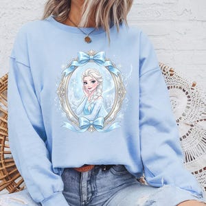 K&ouml;nnte beinhalten: Hellblaues Sweatshirt mit einer gerahmten Illustration einer Frau mit blonden Z&ouml;pfen, die ein blaues Kleid tr&auml;gt. Der Rahmen ist mit blauen Schleifen und B&auml;ndern verziert. Das Sweatshirt hat eine einfarbige hellblaue Farbe.
