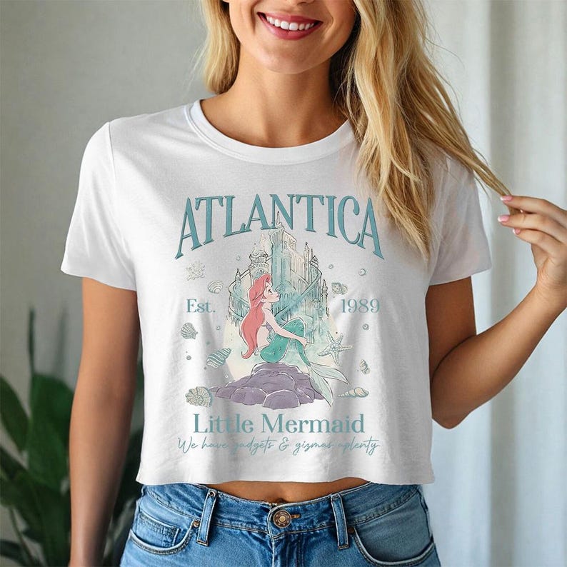 May include: White crop top featuring a teal graphic with the word "Atlantica", the text "Little Mermaid", and the year "1989". The graphic includes a mermaid, a castle, and seashells. The shirt is paired with blue jeans.