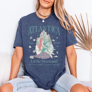 May include: A navy blue t-shirt featuring a graphic of a mermaid with red hair and a green tail, sitting on a rock. The text "Atlantica" arches above the image, with "Little Mermaid" below. The shirt also includes the text "Est. 1989".