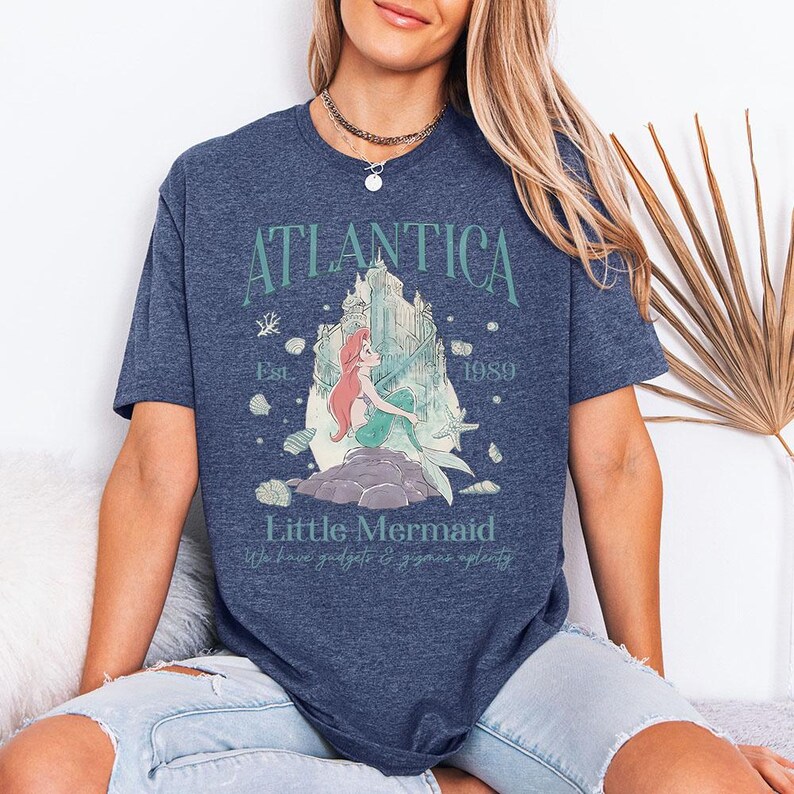 May include: A navy blue t-shirt featuring a graphic of a mermaid with red hair and a green tail, sitting on a rock. The text "Atlantica" arches above the image, with "Little Mermaid" below. The shirt also includes the text "Est. 1989".