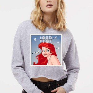 Pode incluir: Um moletom cinza claro com um gr&aacute;fico de Ariel de A Pequena Sereia. O gr&aacute;fico inclui o texto "1989 Ariel's Version" com a imagem de Ariel e gaivotas. O moletom &eacute; um estilo casual.