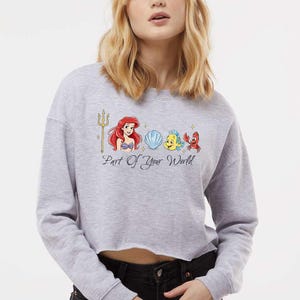 May include: A pale grey cropped sweatshirt with a colourful graphic of Ariel, Flounder, Sebastian, and a seashell. The phrase "Part Of Your World" is printed below the characters. The sweatshirt has long sleeves and a raw hem.