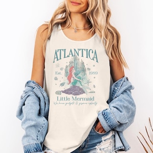 Op de afbeelding: Cr&egrave;mekleurige tanktop met de tekst "Atlantica" boven een afbeelding van een zeemeermin en kasteel. Het ontwerp bevat de tekst "Est. 1989" en "Little Mermaid". De tanktop is mouwloos en heeft een ronde halslijn.