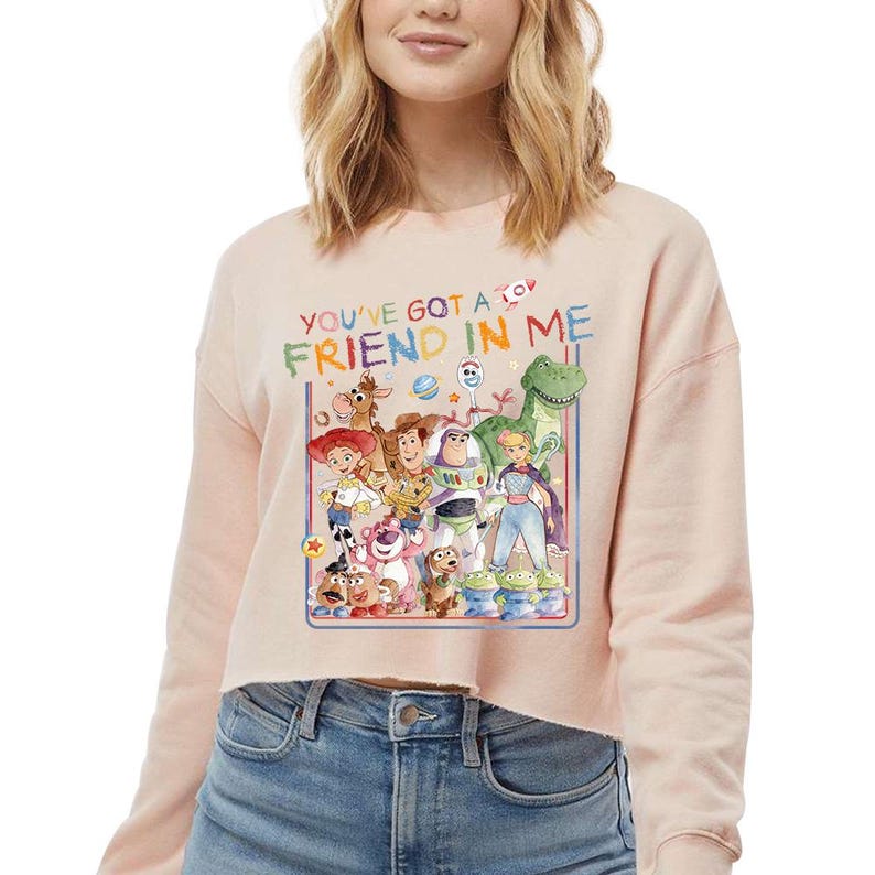 May include: A light pink cropped sweatshirt featuring a colorful graphic of characters from the movie "Toy Story." The graphic includes the text "YOU'VE GOT A FRIEND IN ME." The sweatshirt has long sleeves and a crew neck.
