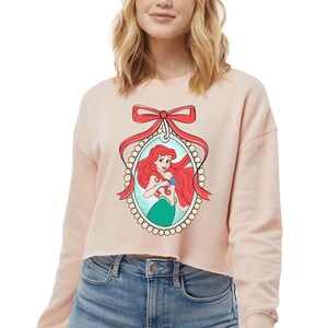 May include: A pale pink cropped sweatshirt with a framed illustration of Ariel from The Little Mermaid. The illustration shows Ariel with red hair and a green tail, framed by a red bow and a pearl-like border.