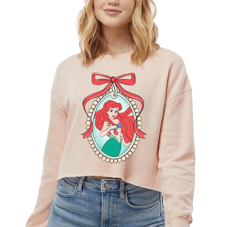 May include: A pale pink cropped sweatshirt with a framed illustration of Ariel from The Little Mermaid. The illustration shows Ariel with red hair and a green tail, framed by a red bow and a pearl-like border.