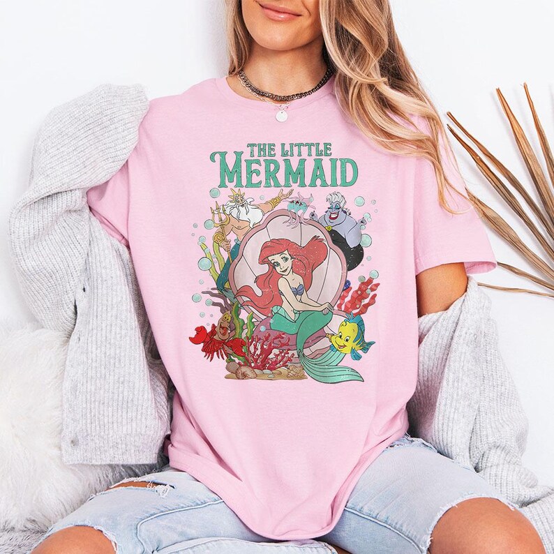 May include: A light pink short-sleeve t-shirt featuring a graphic print of The Little Mermaid characters including Ariel, King Triton, Ursula, Sebastian, and Flounder. The design includes underwater elements like a large seashell and coral.