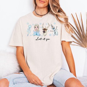 May include: A cream-coloured t-shirt with illustrations of characters from a popular animated film. The design includes a wand, a princess, a reindeer, and a snowman, with the phrase "Let it go" in black script. The shirt has short sleeves and a relaxed fit.