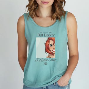May include: A teal tank top with a graphic featuring a cartoon character with red hair and the text "But Daddy I Love Him" in a stylized font. The tank top has a scoop neck and is sleeveless.