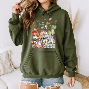 May include: Olive green hooded sweatshirt featuring a colorful graphic of characters from the animated film "Toy Story." The graphic includes the text "You've Got a Friend in Me" and various characters like Woody, Buzz Lightyear, and Rex.
