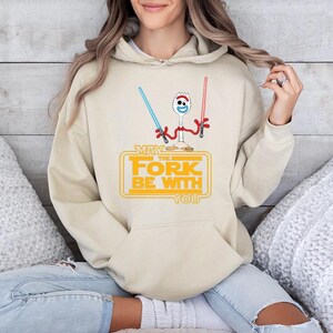May include: A beige hooded sweatshirt featuring a graphic of a white plastic spork character with red pipe cleaner arms, holding a blue and a red lightsaber. Below the character, the yellow text reads: MAY THE FORK BE WITH YOU.
