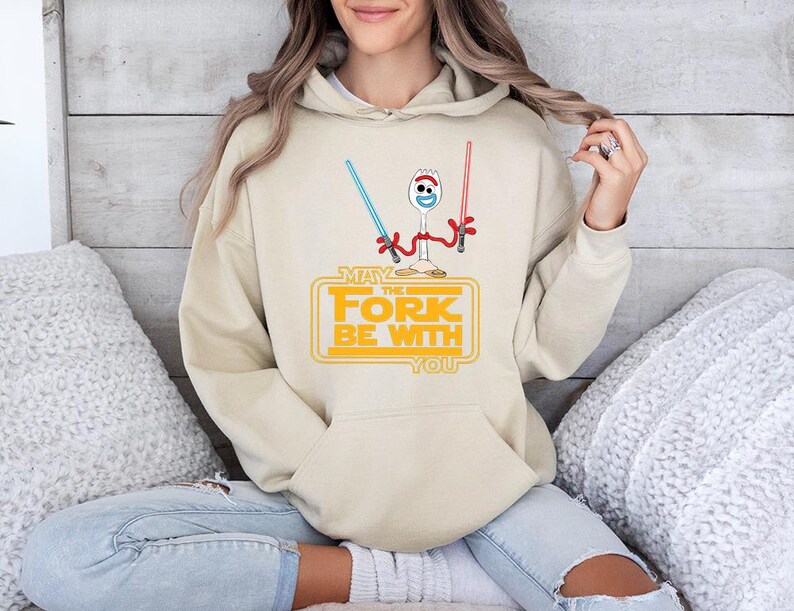 May include: A beige hooded sweatshirt featuring a graphic of a white plastic spork character with red pipe cleaner arms, holding a blue and a red lightsaber. Below the character, the yellow text reads: MAY THE FORK BE WITH YOU.