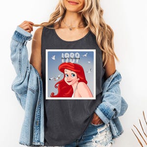 May include: Dark grey tank top featuring a square graphic of Ariel from The Little Mermaid, with red hair and a blue background. The graphic includes the text "1989 Ariel's Version" and images of seagulls.