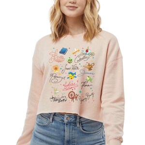 May include: A light peach-coloured cropped sweatshirt featuring a graphic print of various fairy tale icons and character names in cursive script, including Belle, Cinderella, Snow White, Jasmine, Rapunzel, Ariel, Fiona, Tinker Bell, and Sleeping Beauty.