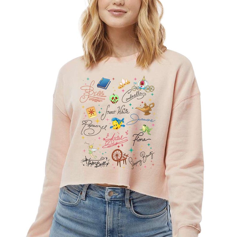 May include: A light peach-coloured cropped sweatshirt featuring a graphic print of various fairy tale icons and character names in cursive script, including Belle, Cinderella, Snow White, Jasmine, Rapunzel, Ariel, Fiona, Tinker Bell, and Sleeping Beauty.