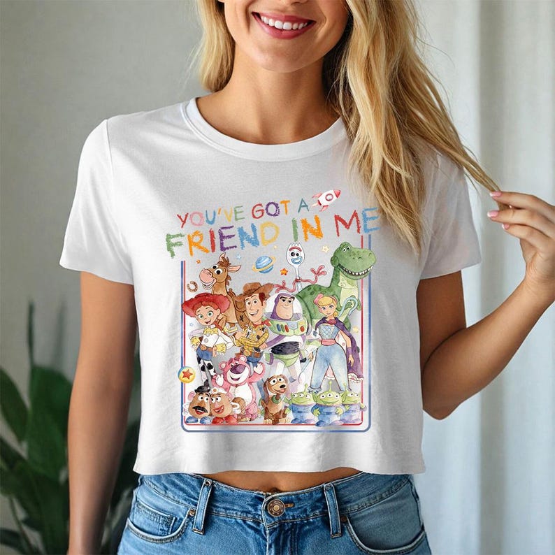 May include: White crop top with a colourful graphic of Toy Story characters and the text "YOU'VE GOT A FRIEND IN ME". The shirt is worn by a person in blue jeans. The image is of a person smiling.
