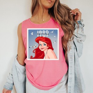May include: Pink tank top featuring a graphic of Ariel from The Little Mermaid. The graphic includes the text "1989 Ariel's Version" with seagulls in the background. The tank top is sleeveless and made of a soft material.