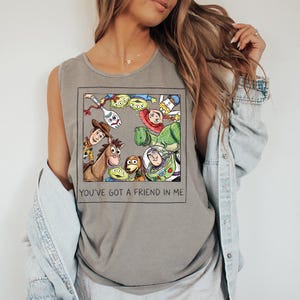 May include: Grey tank top with a framed graphic of Toy Story characters. The image includes Woody, Buzz Lightyear, and other characters from the movie. The text "YOU'VE GOT A FRIEND IN ME" is printed below the graphic.