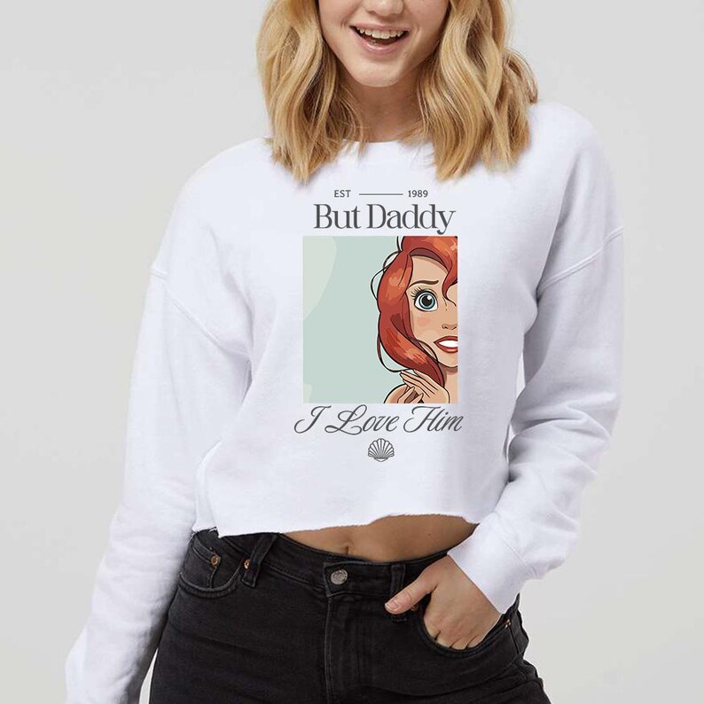 May include: White cropped sweatshirt with the text "But Daddy I Love Him" and an illustration of a red-haired character. The sweatshirt has a crew neck and long sleeves. The illustration is in a rectangular box.