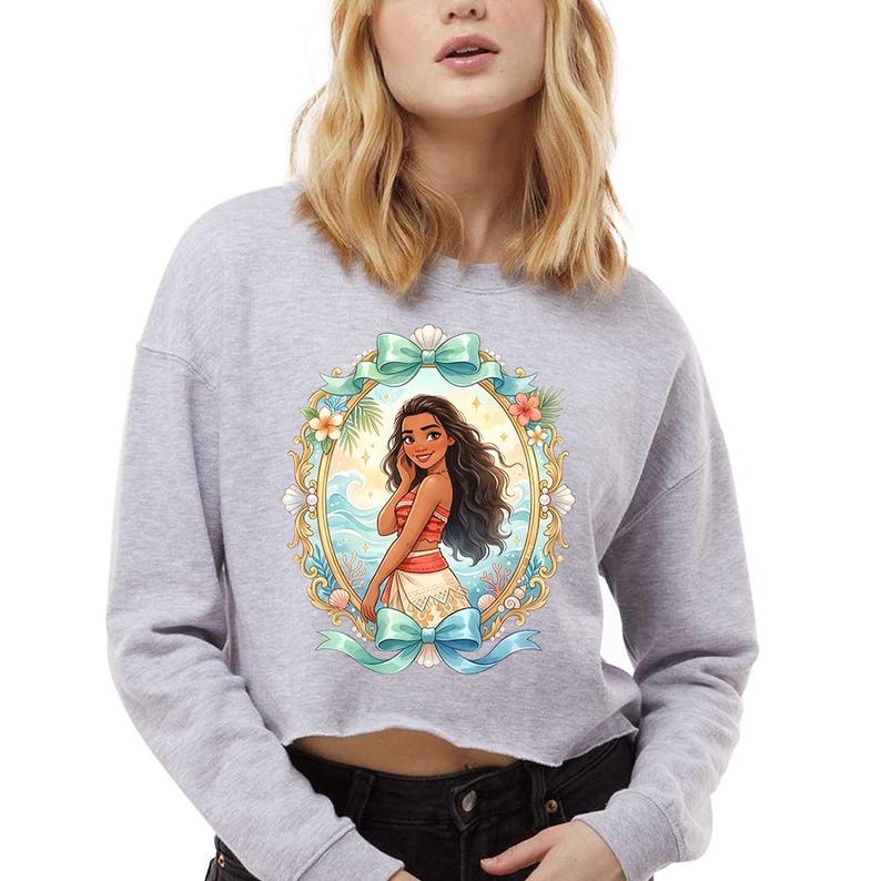 Peut inclure: Un sweat-shirt gris court avec une illustration color&eacute;e d'un personnage de dessin anim&eacute; dans un cadre ovale. Le cadre comprend un n&oelig;ud bleu sarcelle, des fleurs et des coquillages. Le personnage a de longs cheveux fonc&eacute;s et porte une tenue rouge et blanche.