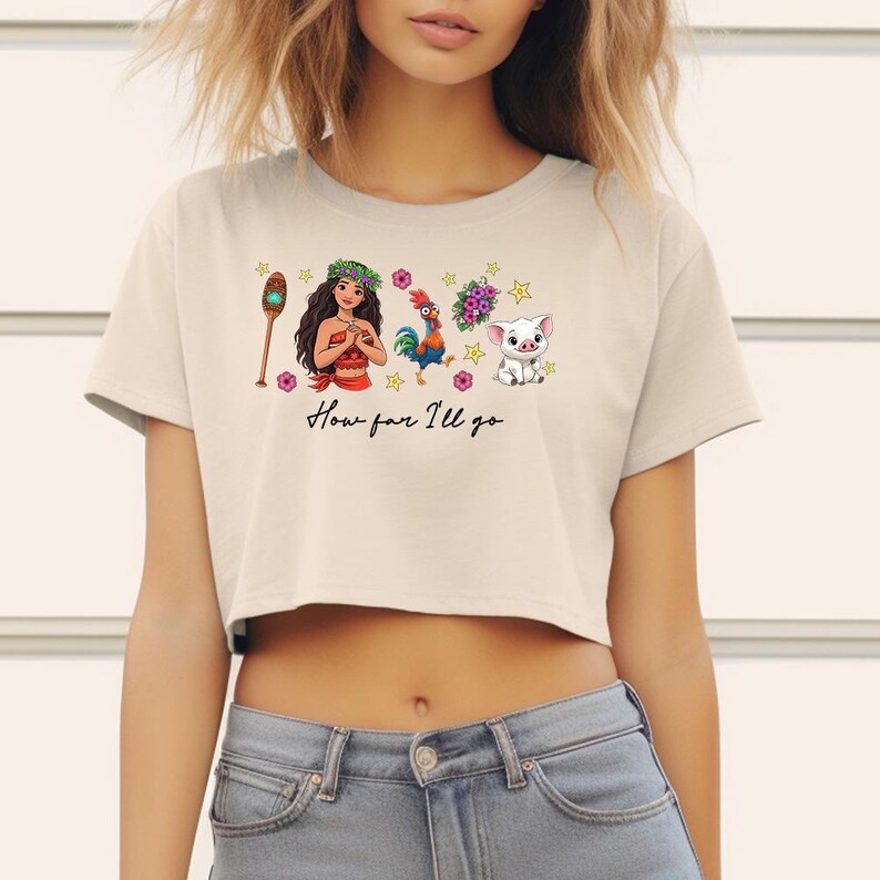 May include: Cream-coloured crop top with a colourful graphic design featuring characters and the text "How far I'll go". The design includes a woman, rooster, pig, and other elements. The shirt is made of a soft material.