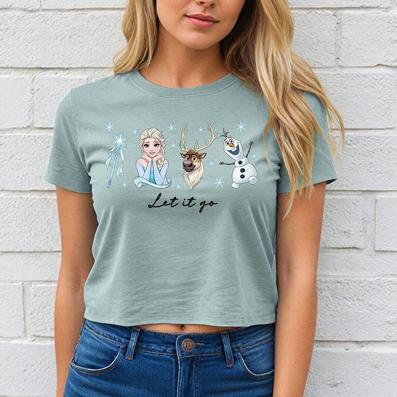 May include: A light green crop top featuring Elsa, Sven, and Olaf from Frozen. The shirt has the phrase "Let it go" in a script font. The design includes snowflakes and is set against a white brick wall.