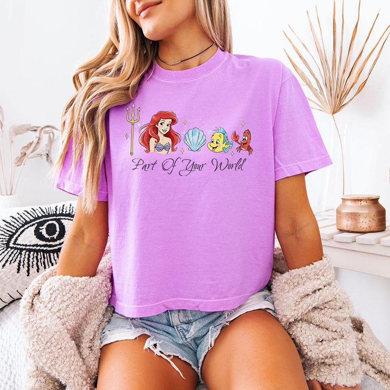 May include: A lilac-colored t-shirt featuring a graphic of Ariel, a seashell, Flounder, and Sebastian. The phrase "Part of Your World" is printed below the characters. The shirt is a short-sleeved, crew neck style.