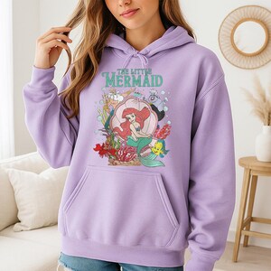 May include: A light purple hooded sweatshirt featuring a graphic print of The Little Mermaid characters including Ariel, King Triton, Ursula, Sebastian, and Flounder. The design includes a large pink seashell, coral, and bubbles on the front.