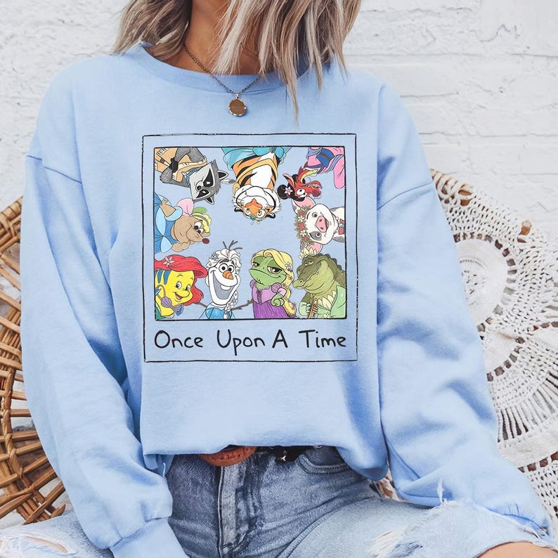 May include: Light blue sweatshirt featuring a framed graphic with cartoon characters and the text "Once Upon A Time." The graphic includes a raccoon, tiger, pig, and other characters. The sweatshirt is a solid light blue color.