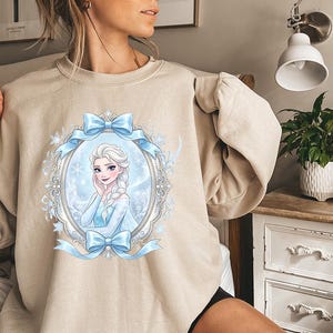 K&ouml;nnte beinhalten: Beiges Sweatshirt mit einer gerahmten Illustration einer Cartoon-Frau mit blonden Z&ouml;pfen, die ein blaues Kleid tr&auml;gt. Der Rahmen ist mit blauen Schleifen und Schneeflocken verziert. Das Sweatshirt hat eine neutrale Farbe.