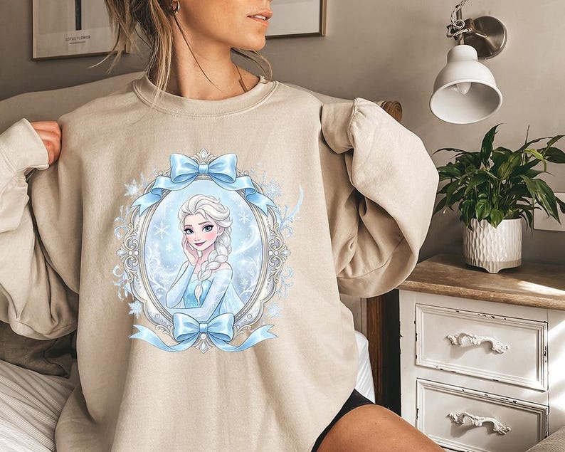 K&ouml;nnte beinhalten: Beiges Sweatshirt mit einer gerahmten Illustration einer Cartoon-Frau mit blonden Z&ouml;pfen, die ein blaues Kleid tr&auml;gt. Der Rahmen ist mit blauen Schleifen und Schneeflocken verziert. Das Sweatshirt hat eine neutrale Farbe.