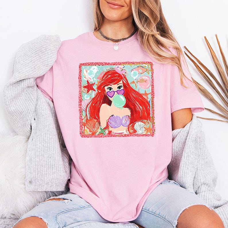 May include: Pink t-shirt featuring a graphic of a red-haired mermaid wearing heart-shaped sunglasses and blowing a bubble. The mermaid is surrounded by seashells and starfish. The shirt is a light pink color.