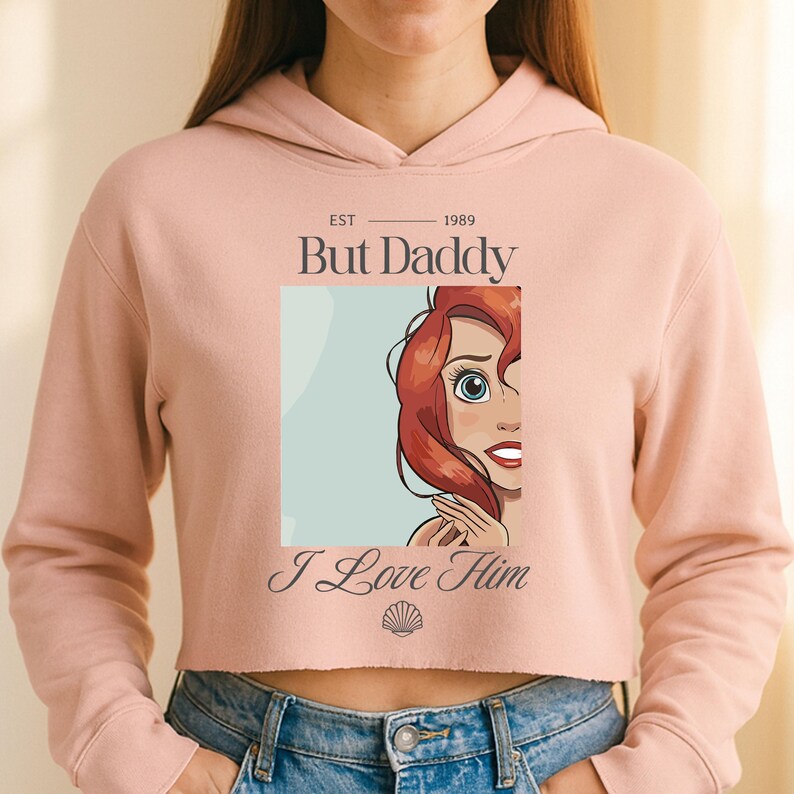 K&ouml;nnte beinhalten: Ein hellrosa Crop-Hoodie mit dem Text "EST 1989 But Daddy I Love Him" und einer Cartoon-Illustration einer Figur mit roten Haaren und blauen Augen. Der Hoodie hat lange &Auml;rmel und eine Kapuze mit Kordelzug.