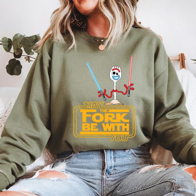 May include: An olive green crewneck sweatshirt featuring a graphic of a white spork character holding a blue lightsaber and a red lightsaber. Below the character, the yellow text reads, "MAY THE FORK BE WITH YOU" in a bold, blocky font style.